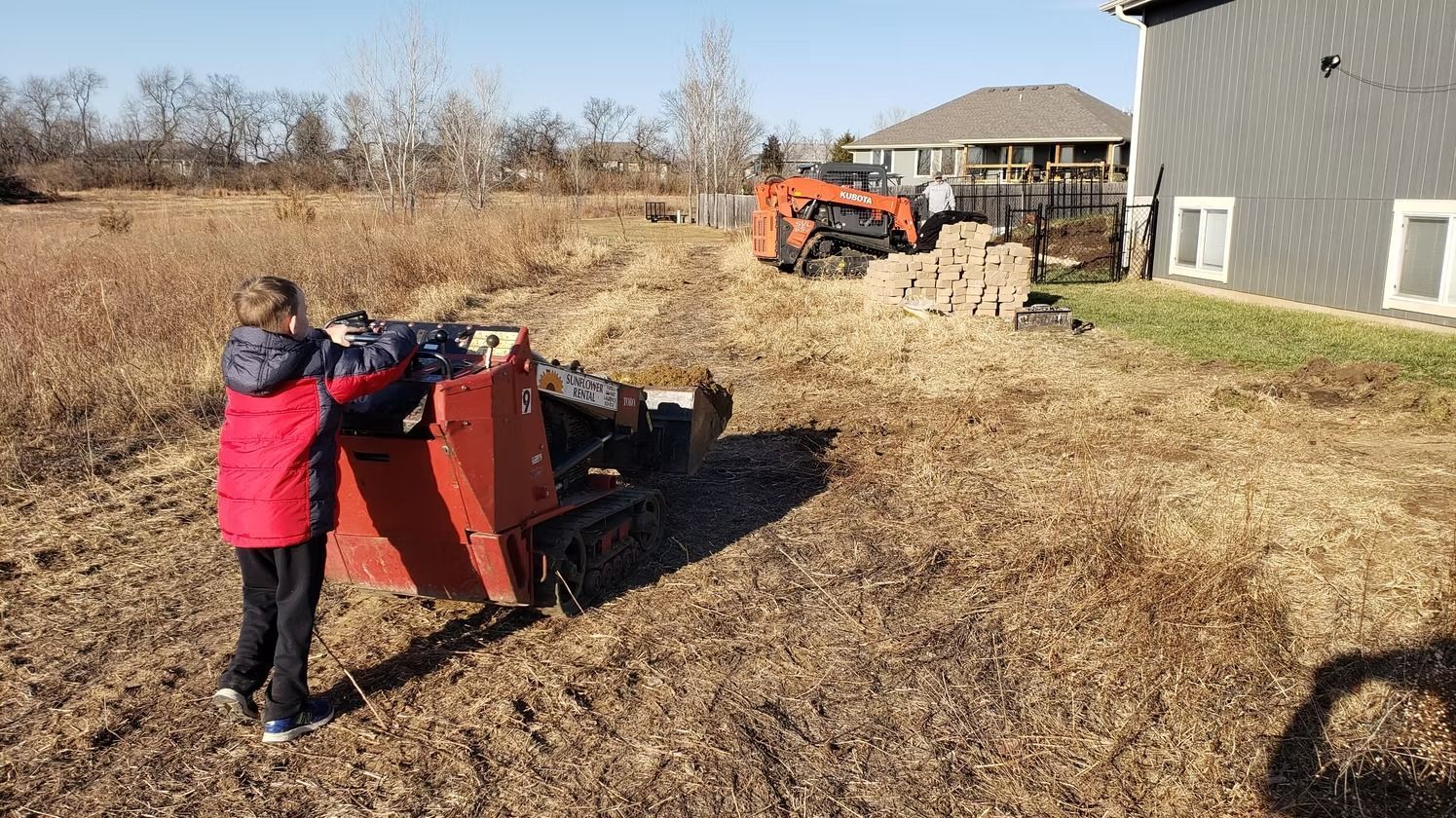 Person operating an orange mini skid steer on a grassy lot next to a house. A larger excavator and brick pile are in the background.