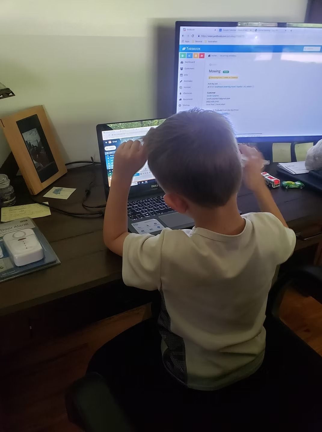 Boy seated at desk, looking at laptop and monitor screens. Hands raised, possibly stretching.