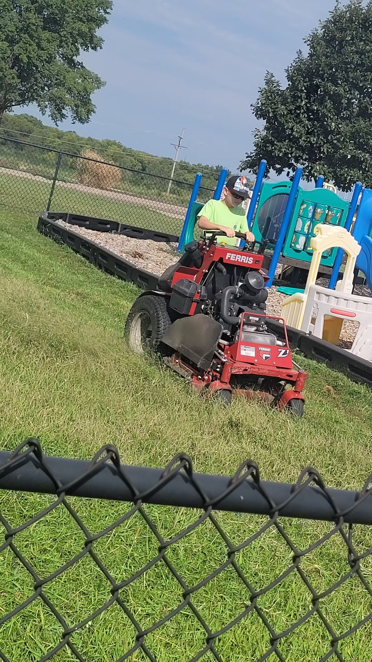 Person mowing grass near a playground with a red riding mower.