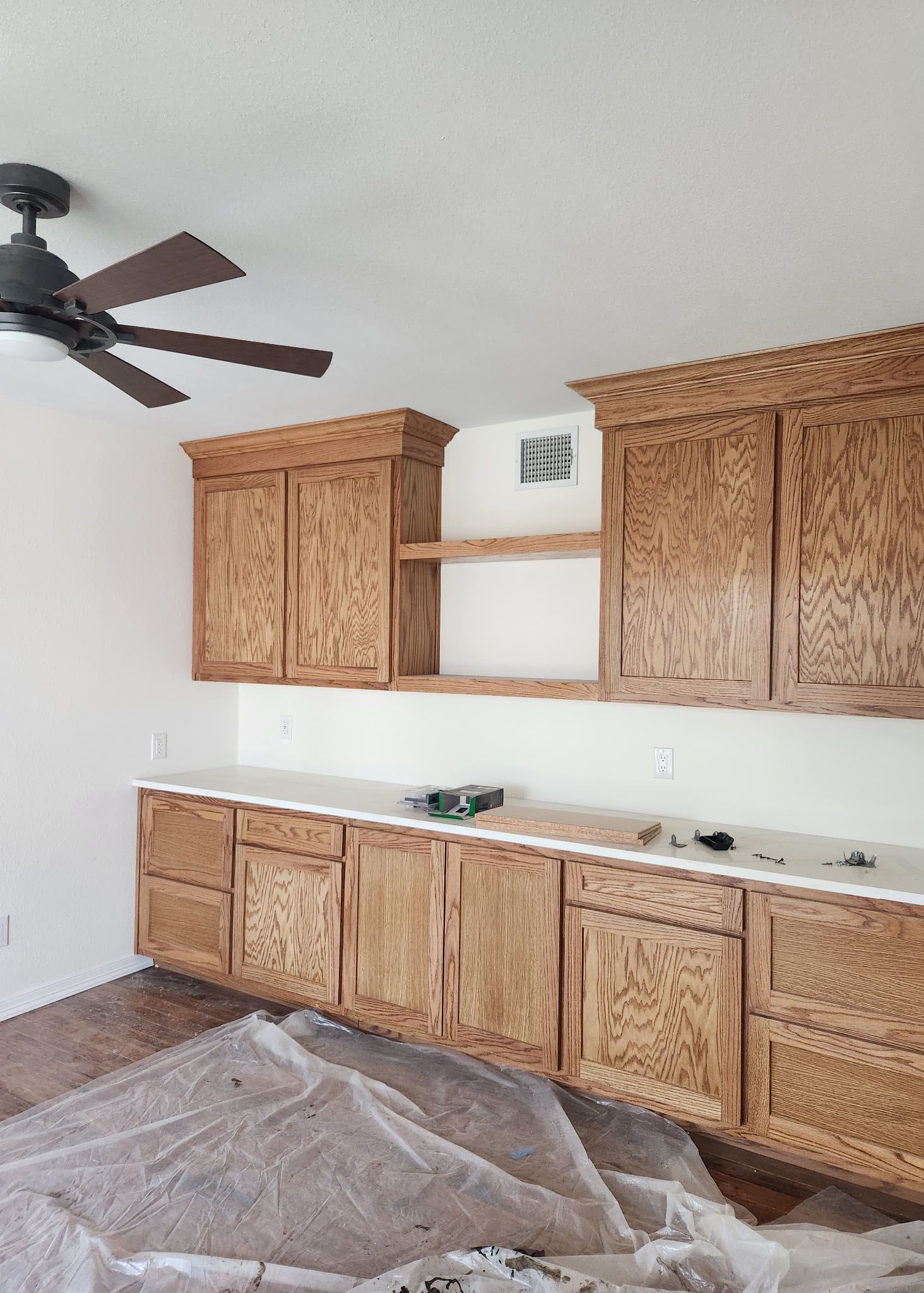 Wooden cabinets with white countertop and shelving, under a ceiling fan in a room.