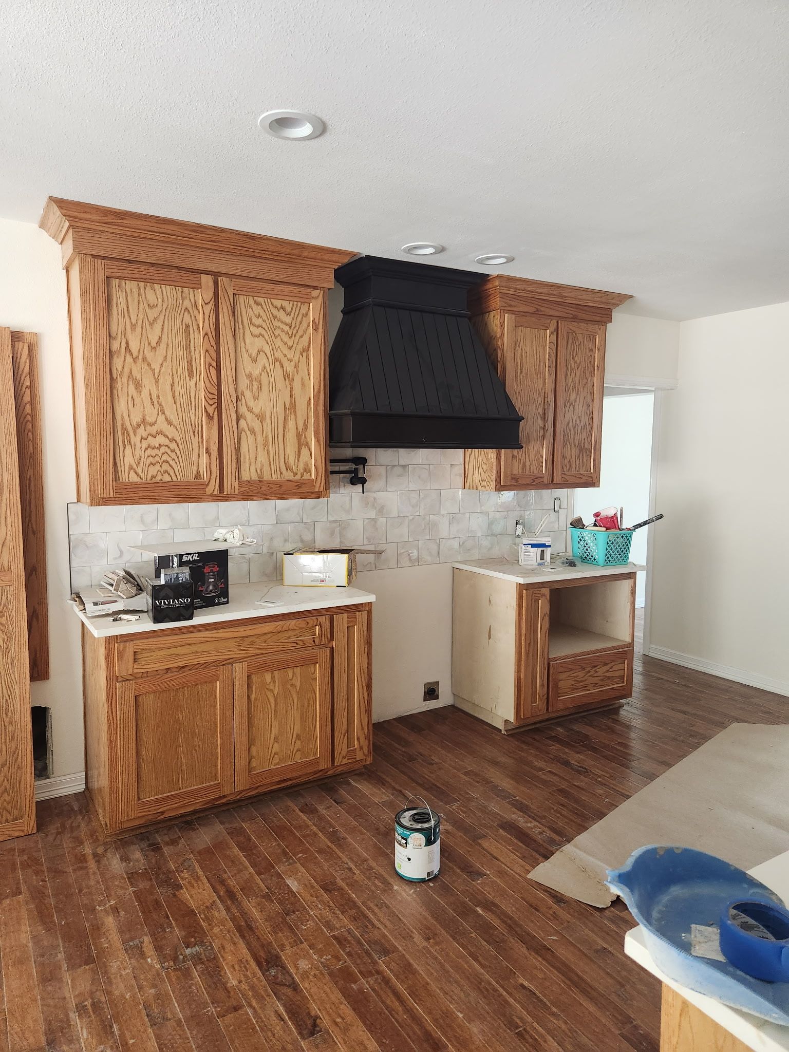Kitchen under construction: wood cabinets, black range hood, white tile backsplash, hardwood floor.