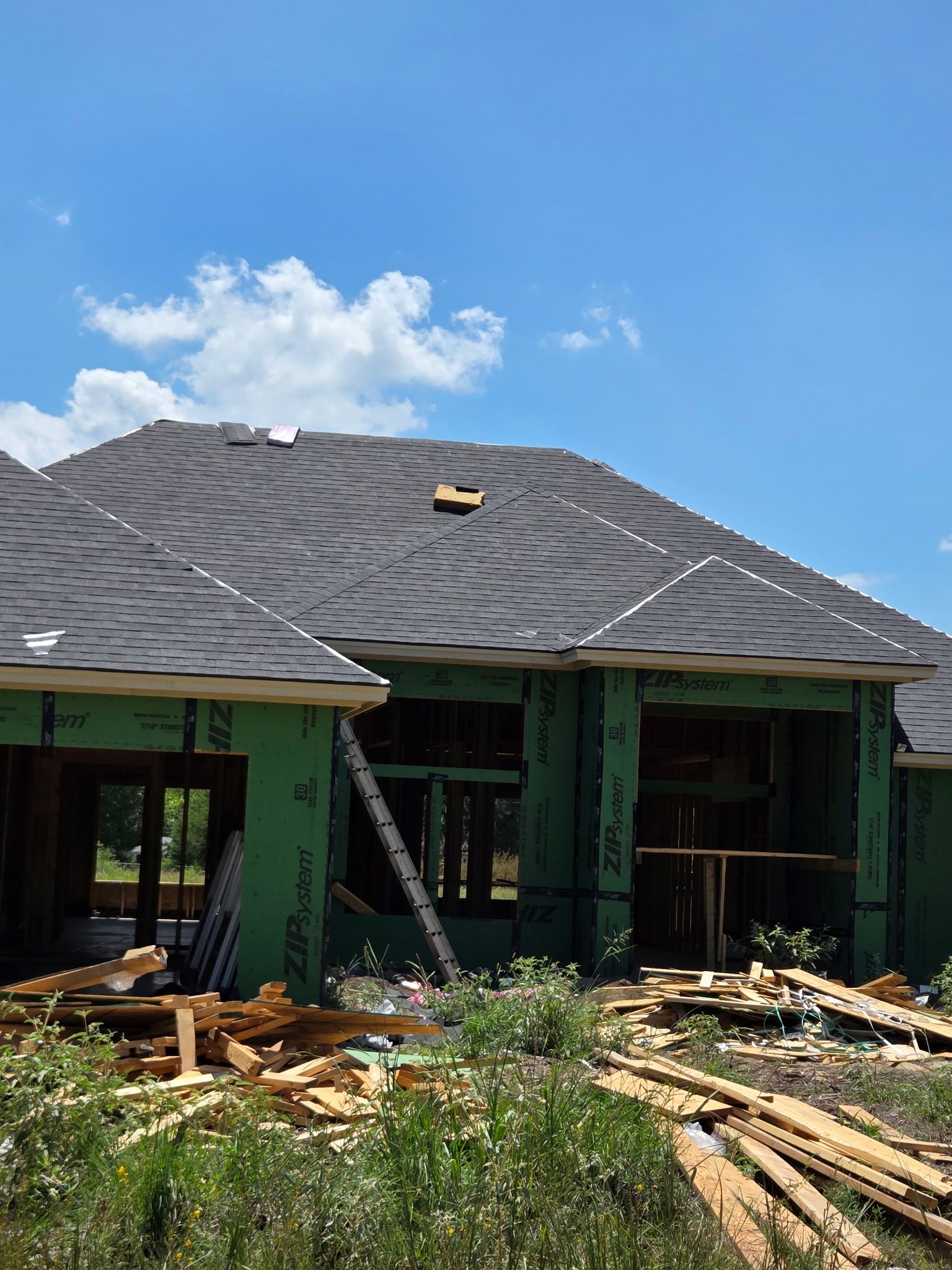 House under construction; roof with shingles, green wrap, blue sky, and wood debris.