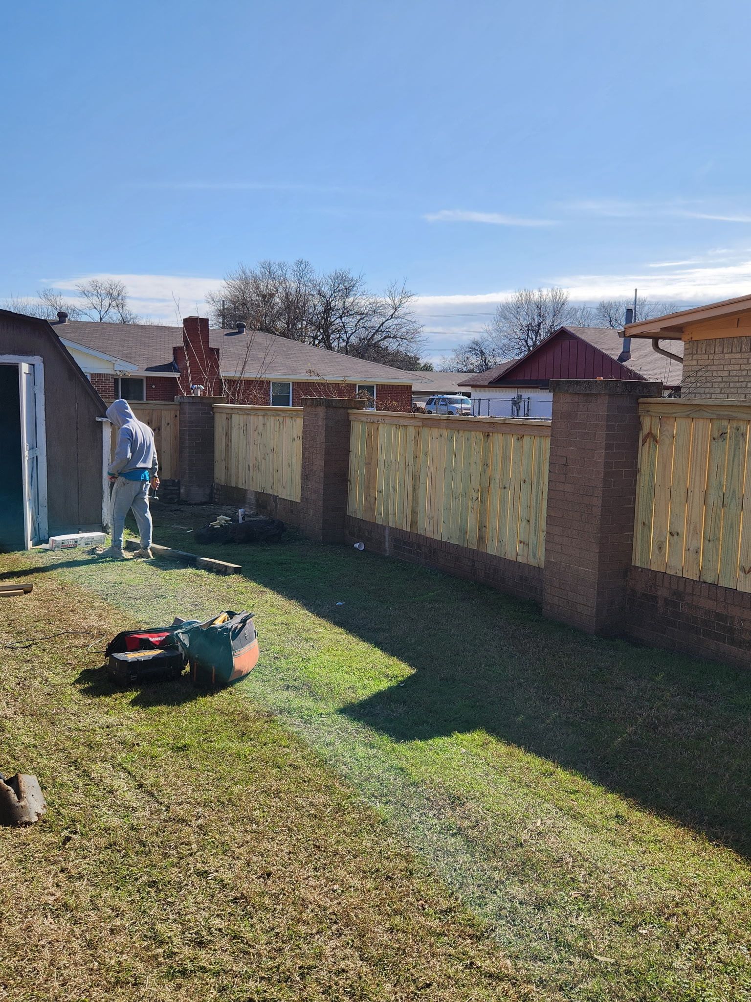 Fence being built, brick pillars, wood panels, person in protective suit spraying; sunny day.