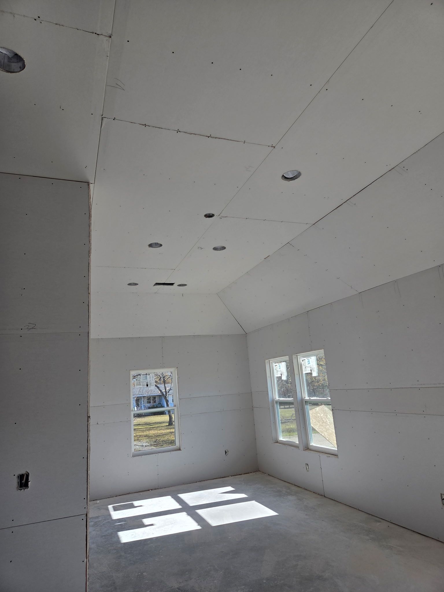 Interior of a room under construction with drywall and recessed lighting. Windows let in sunlight.