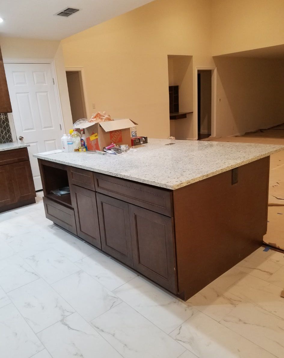 Kitchen island with granite countertop and dark wood cabinets. Boxes and items on the countertop. Light floor and yellow walls.