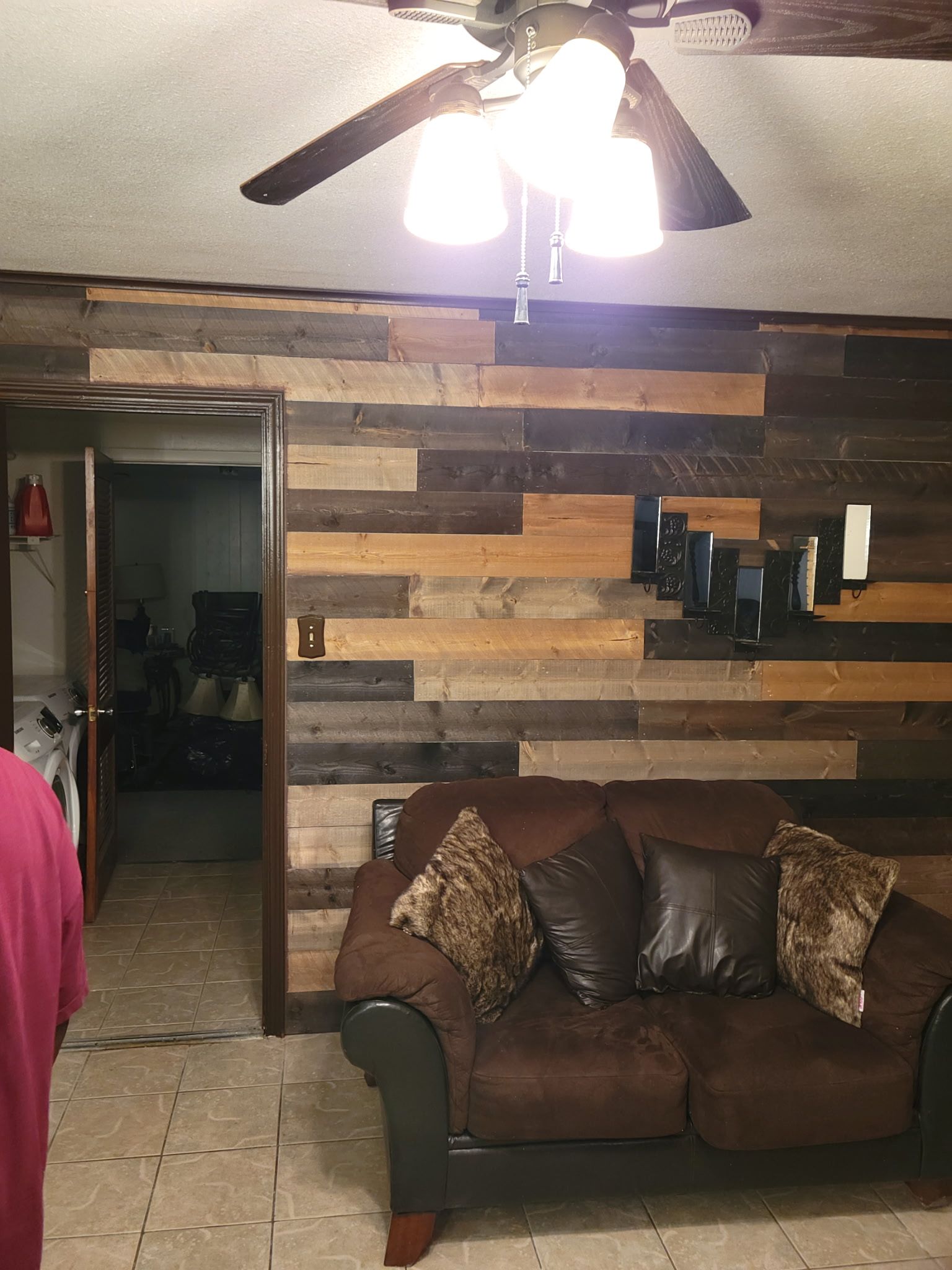 Living room with wood-paneled wall, brown couch with pillows, ceiling fan, and open doorway to another room.