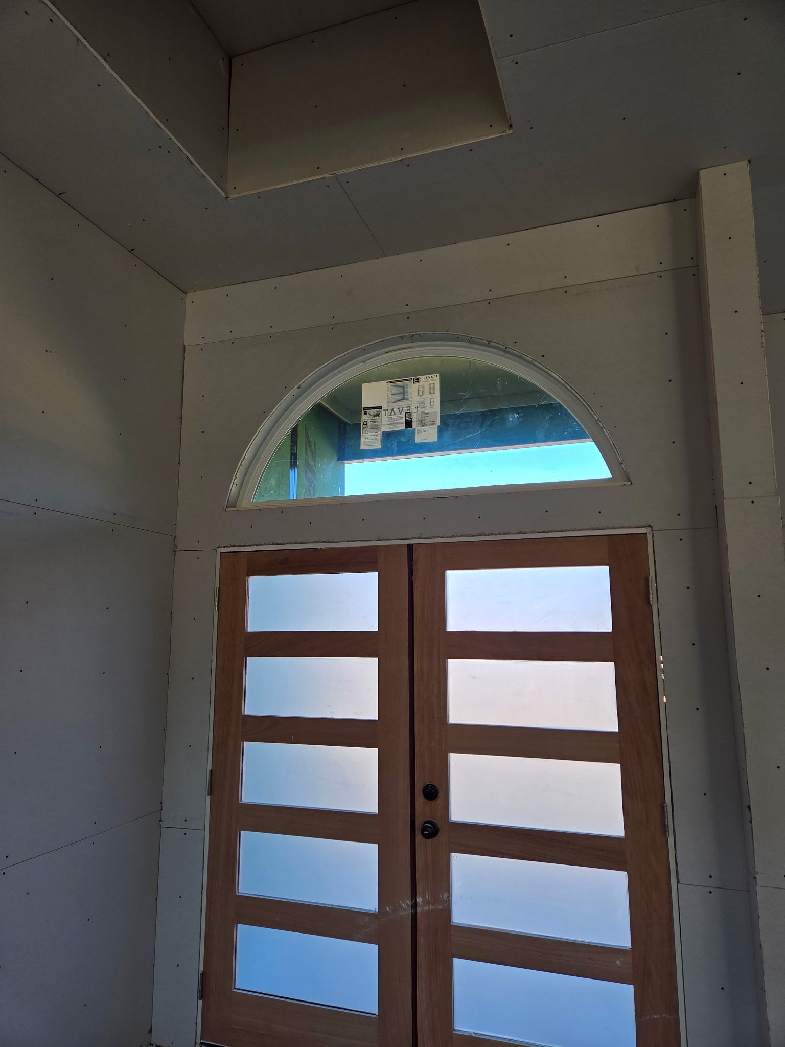 Double wooden doors with frosted glass panels, topped by an arched window. Drywall surrounds the doorway.