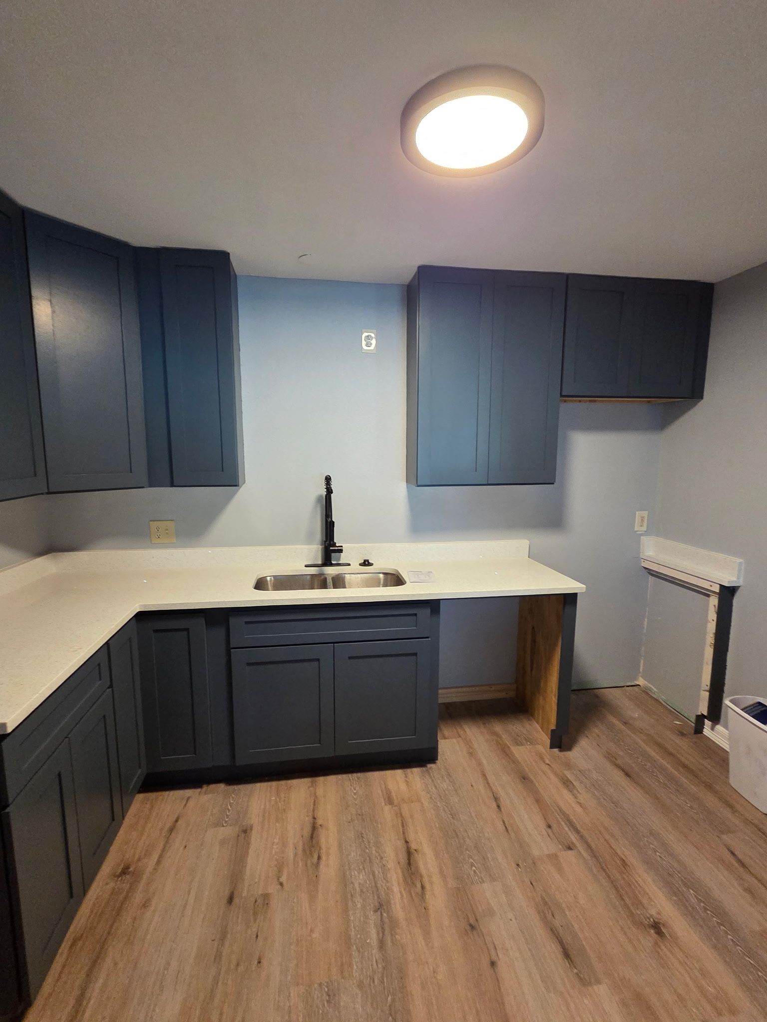 Blue kitchen cabinets, white countertop, stainless steel sink, wood floor, and a light fixture.