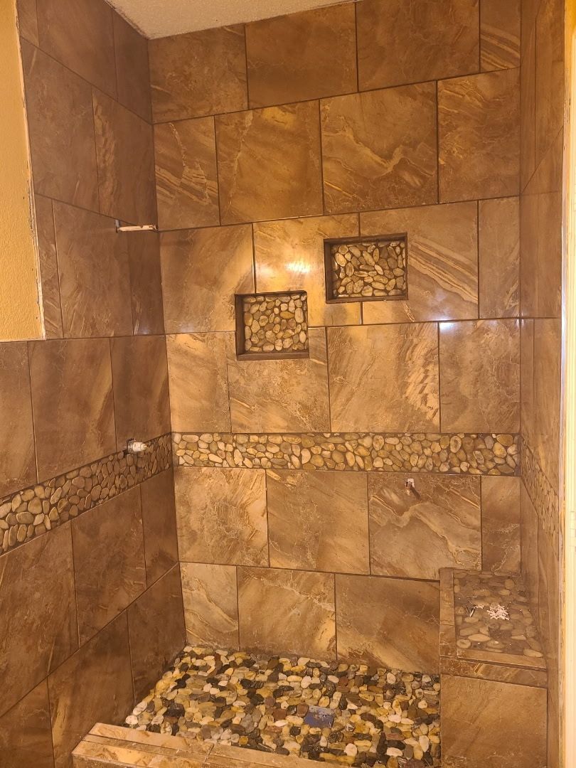 Tiled shower interior with brown, stone-like tiles, two decorative niches, and a pebble-stone floor.