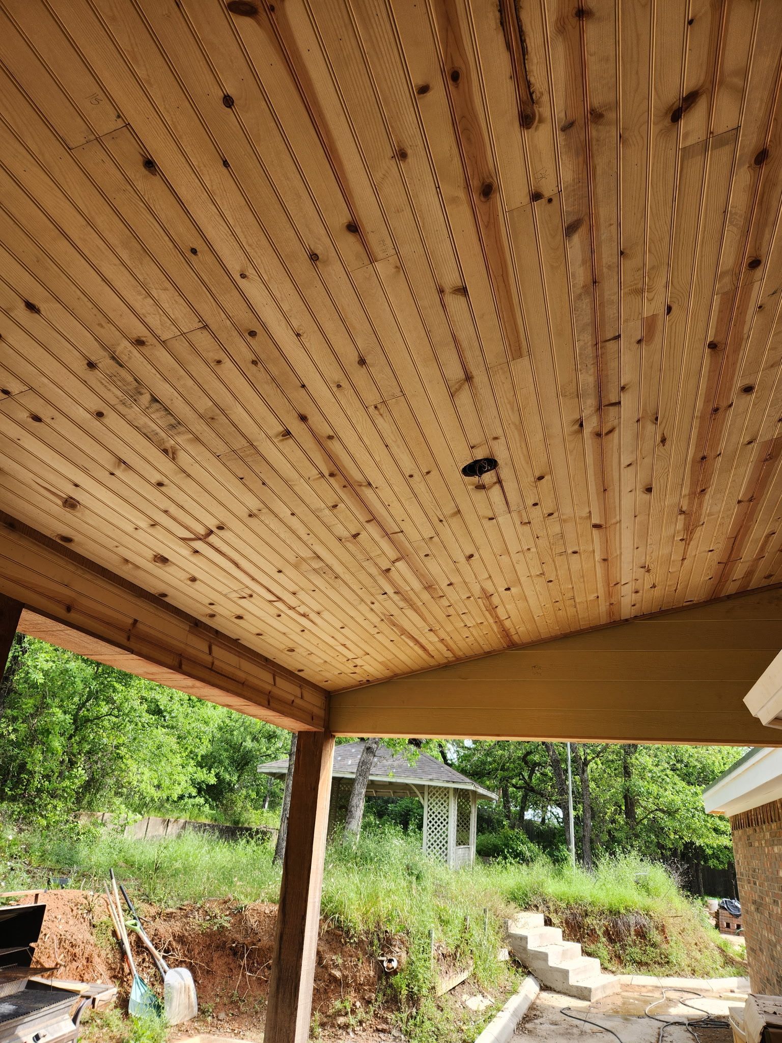 Wooden porch ceiling with drilled ventilation holes, view of greenery and a gazebo in the background.