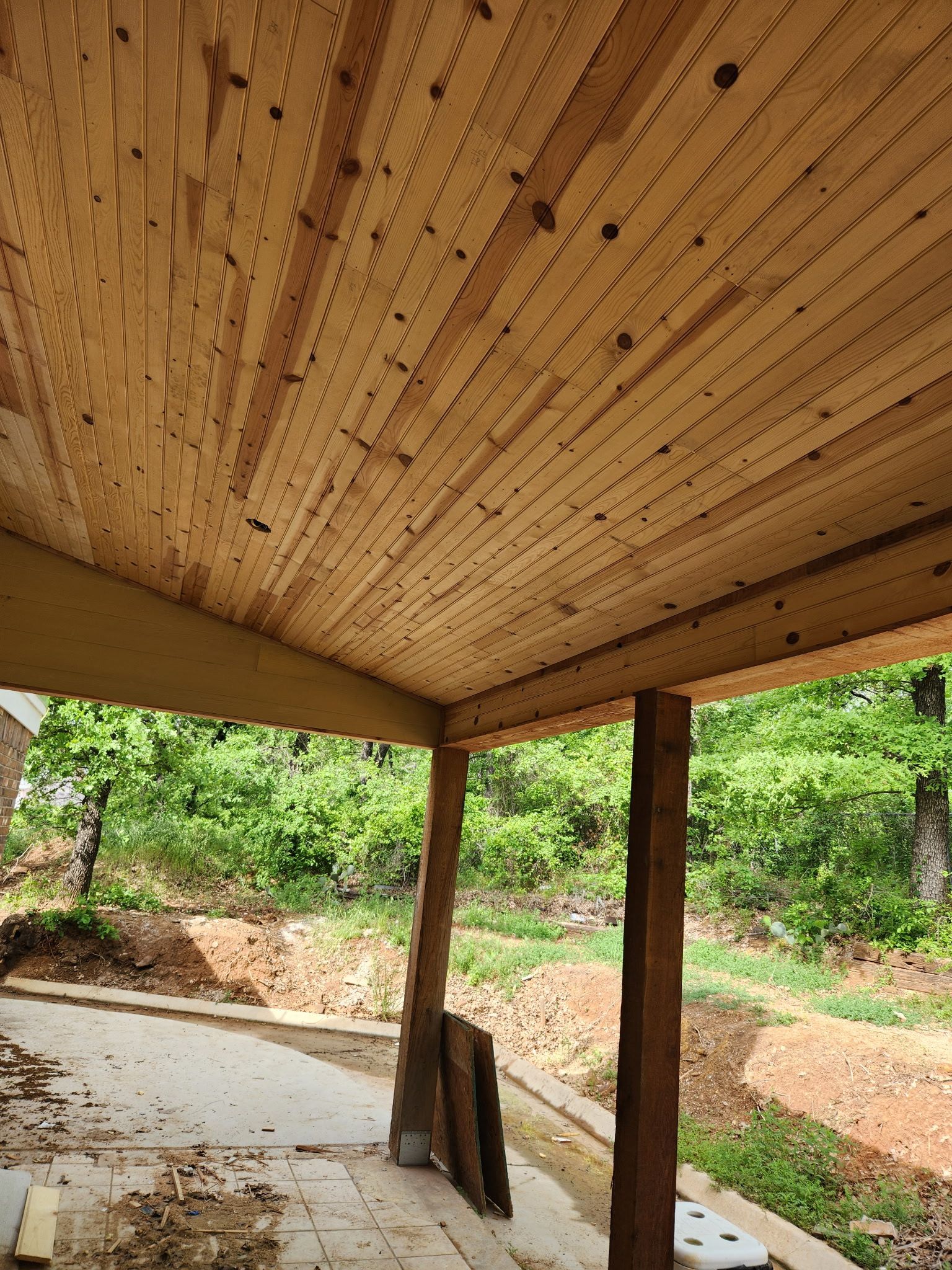 Wooden porch ceiling with exposed beams and support posts overlooking a wooded area.