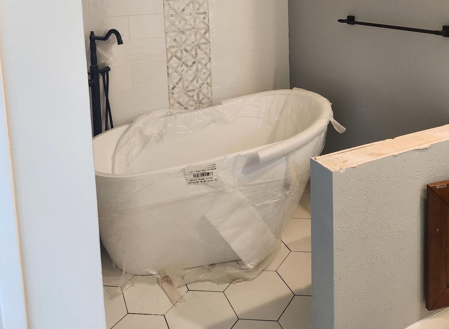 White oval bathtub in a bathroom, plastic wrap, hexagon tile floor, black faucet.