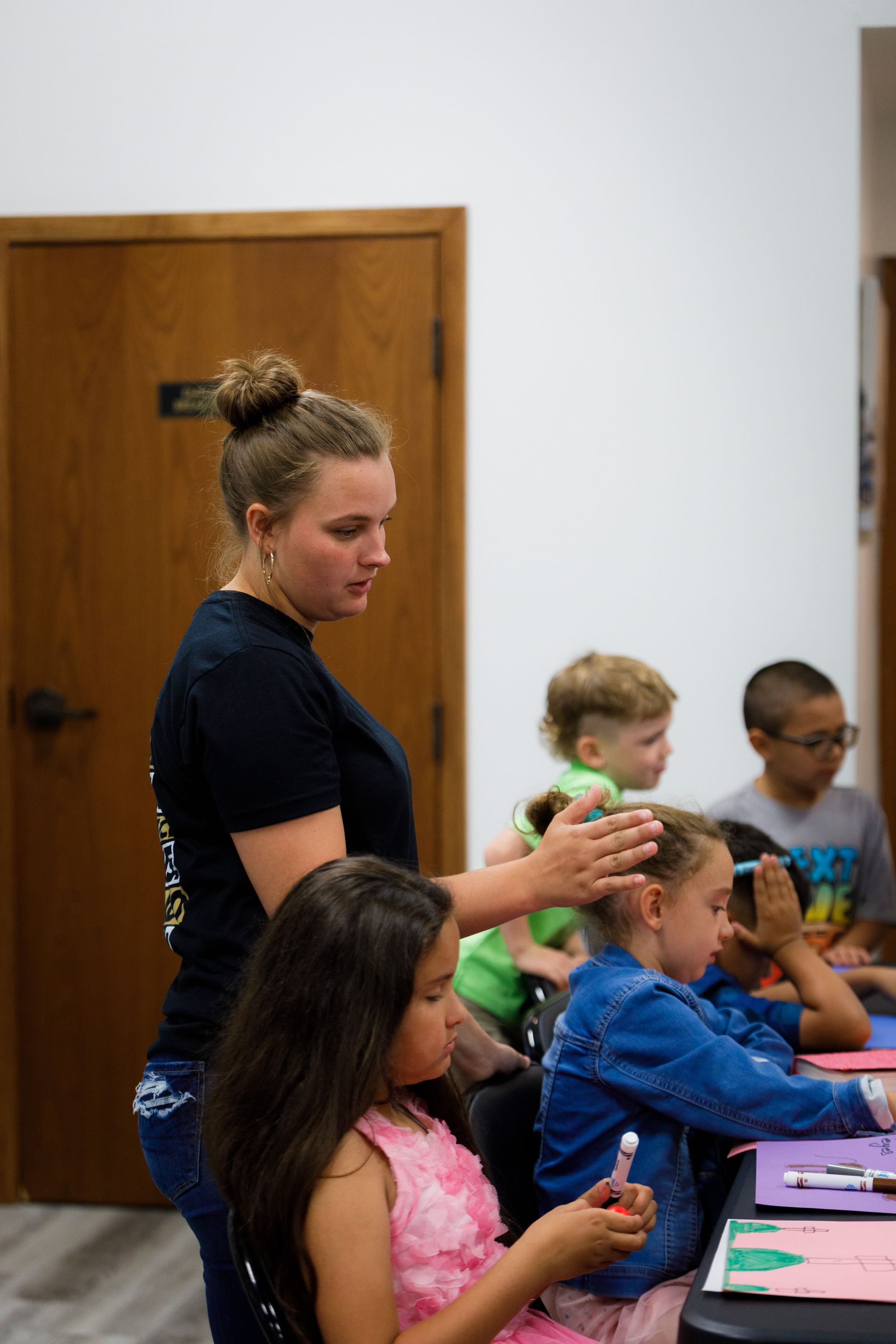 Woman assisting children at a table. Classroom setting, with children coloring.