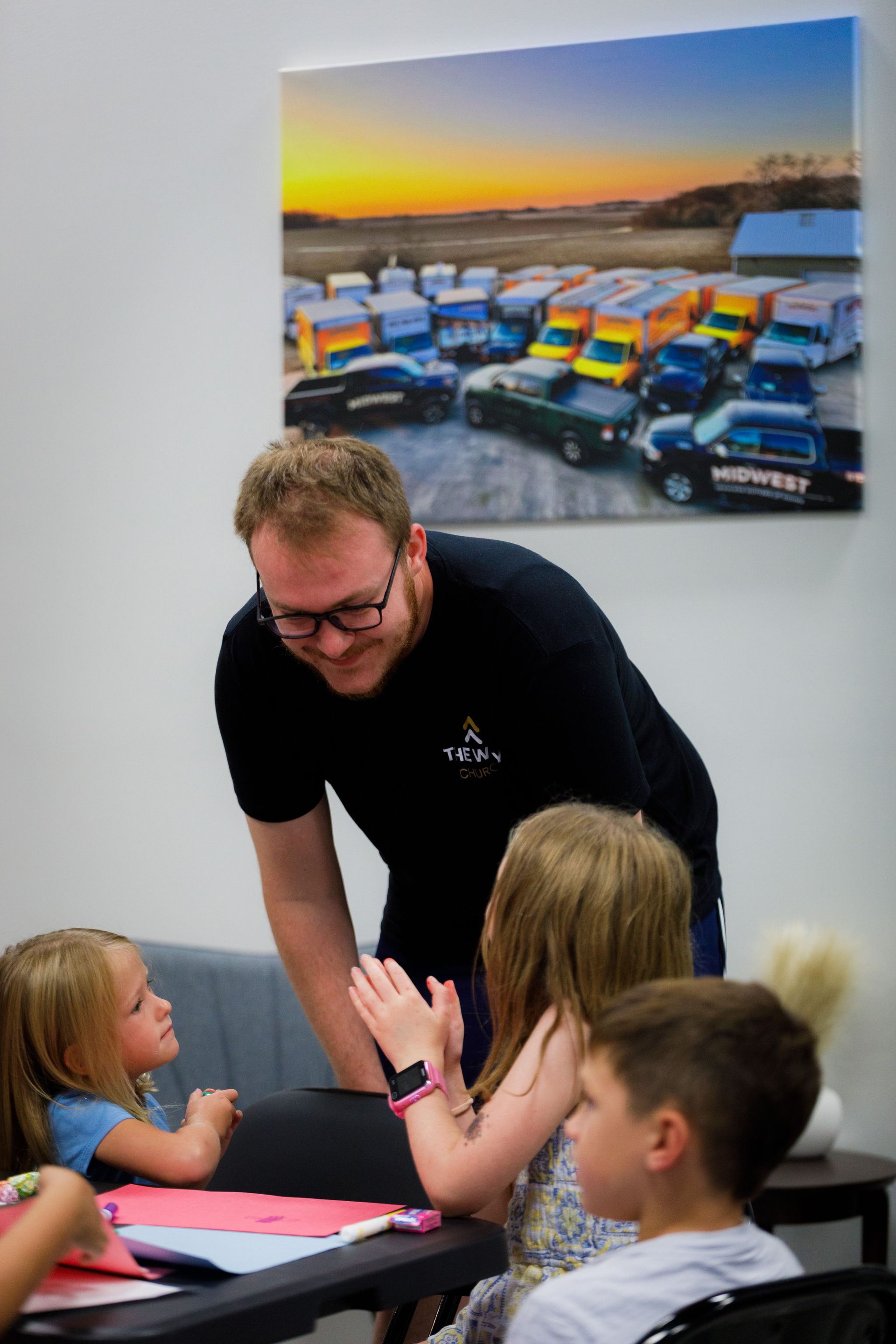 Man smiles at children seated at a table, painting. A photo of vehicles hangs on the wall.