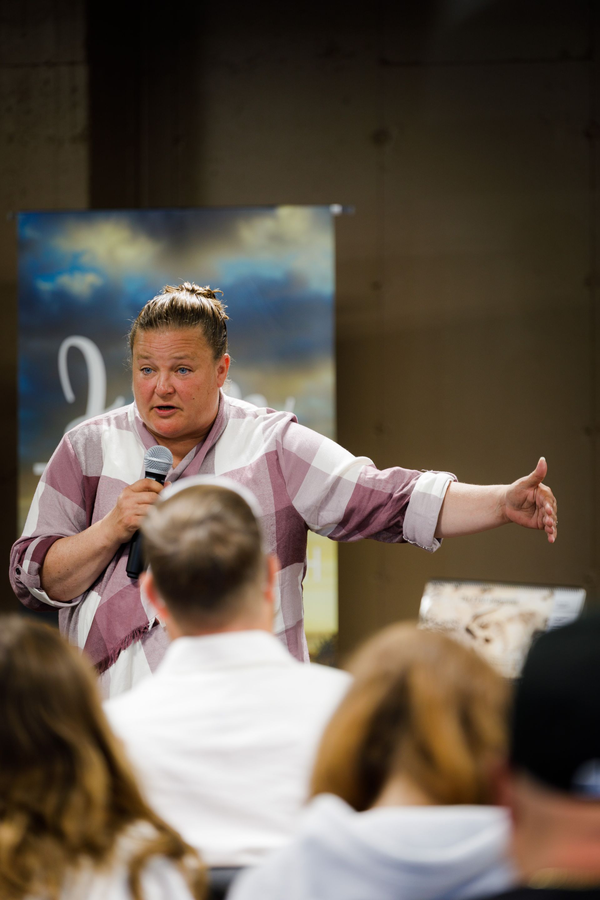 Woman speaking into a microphone, gesturing to an audience. Plaid shirt, bun. Indoor setting, blue backdrop.