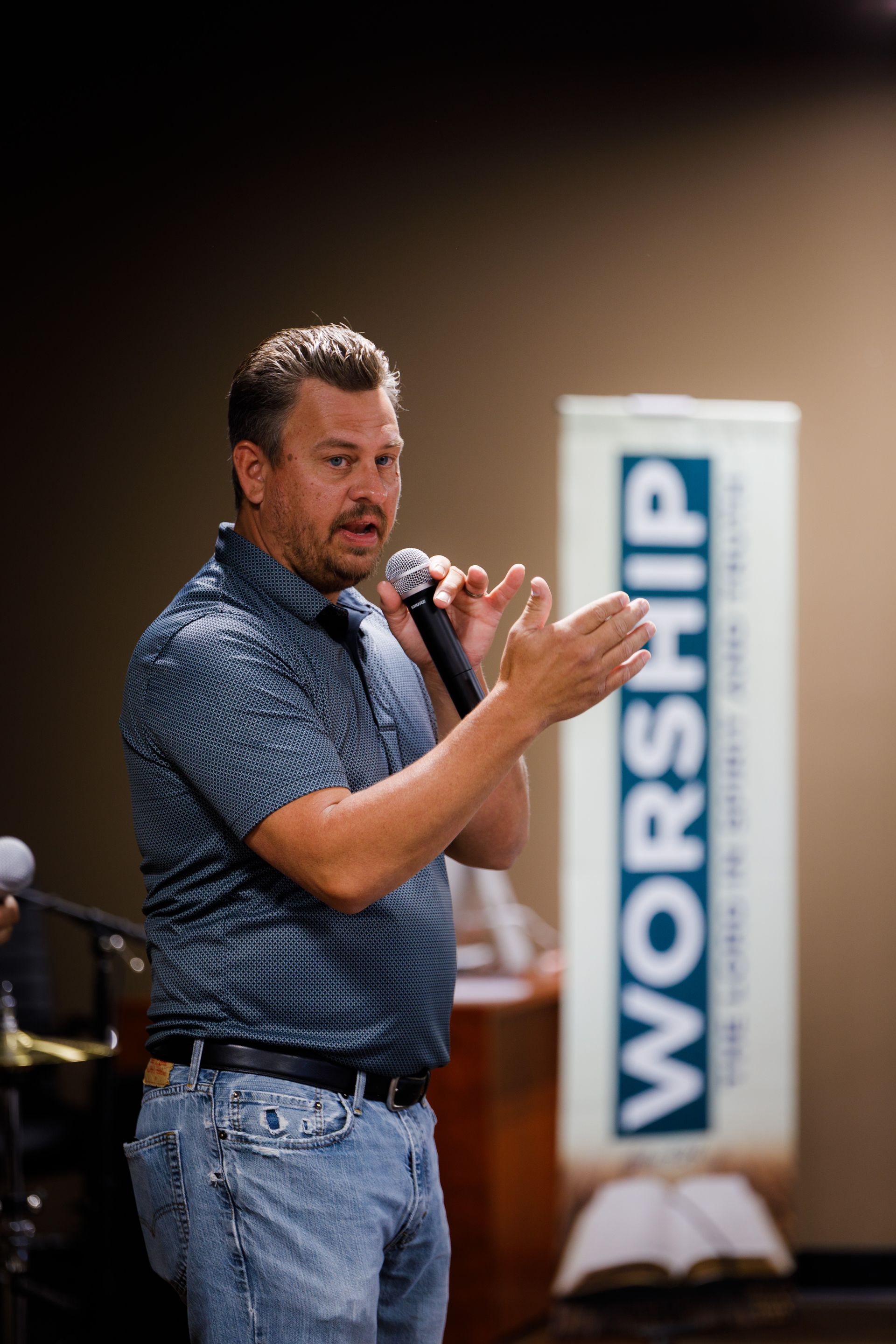 Man with microphone speaking, gesturing in front of a banner that says Worship.