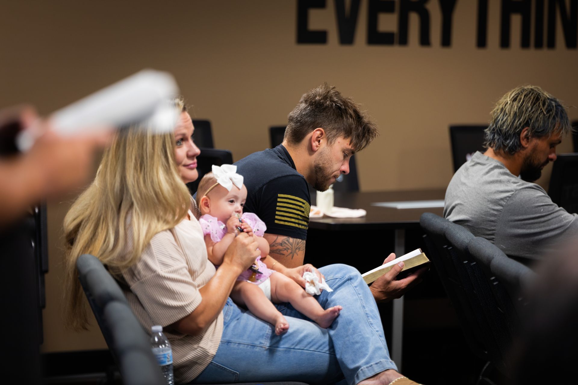 A family sits together in a room: mom, dad reading, and baby. Everyone's seated, light-colored walls, neutral tones.