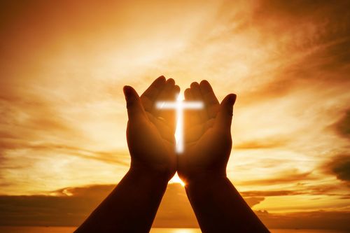 A person is holding a luminated cross in their hands in front of a sunset.