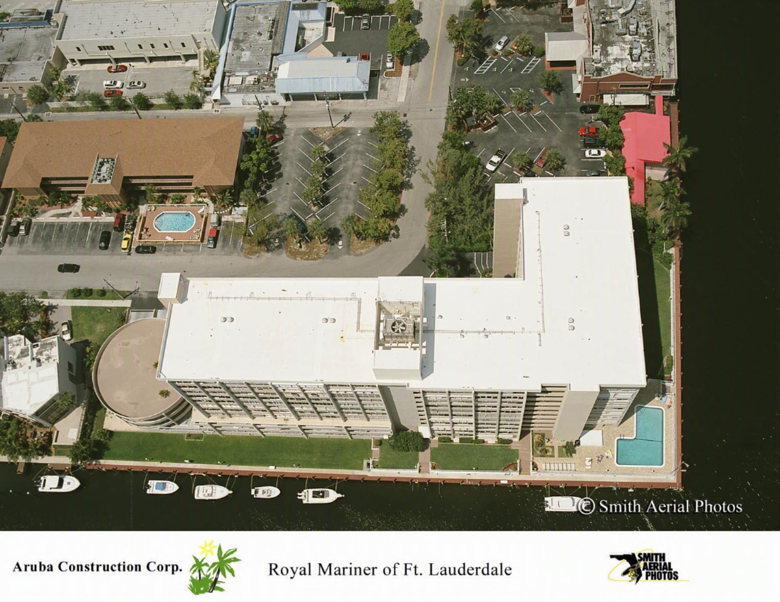 Aerial view of a white waterfront building with pool and parked cars beside a marina.