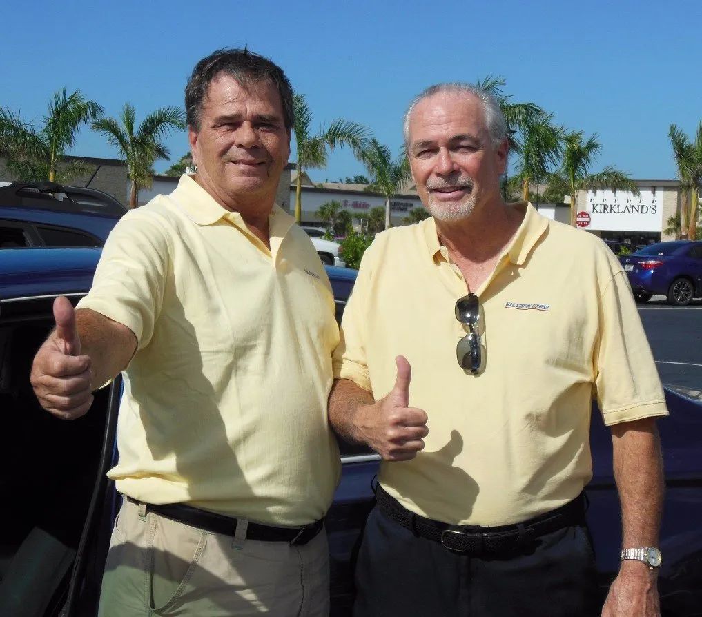 Two men in yellow shirts give thumbs-up outside a shopping area on a sunny day.