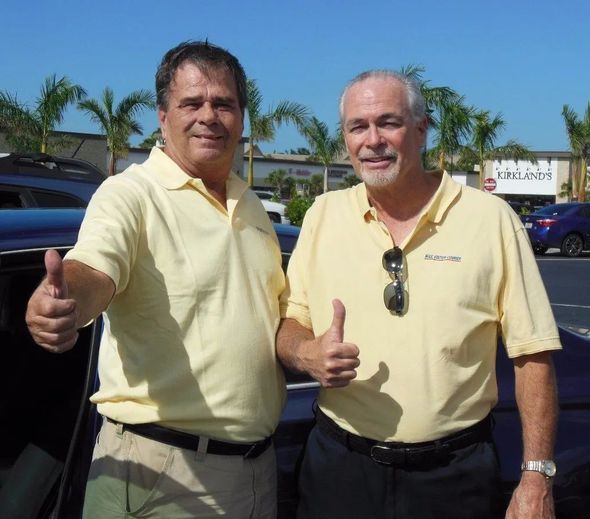 Two men in yellow shirts give thumbs-up outside a shopping area on a sunny day.