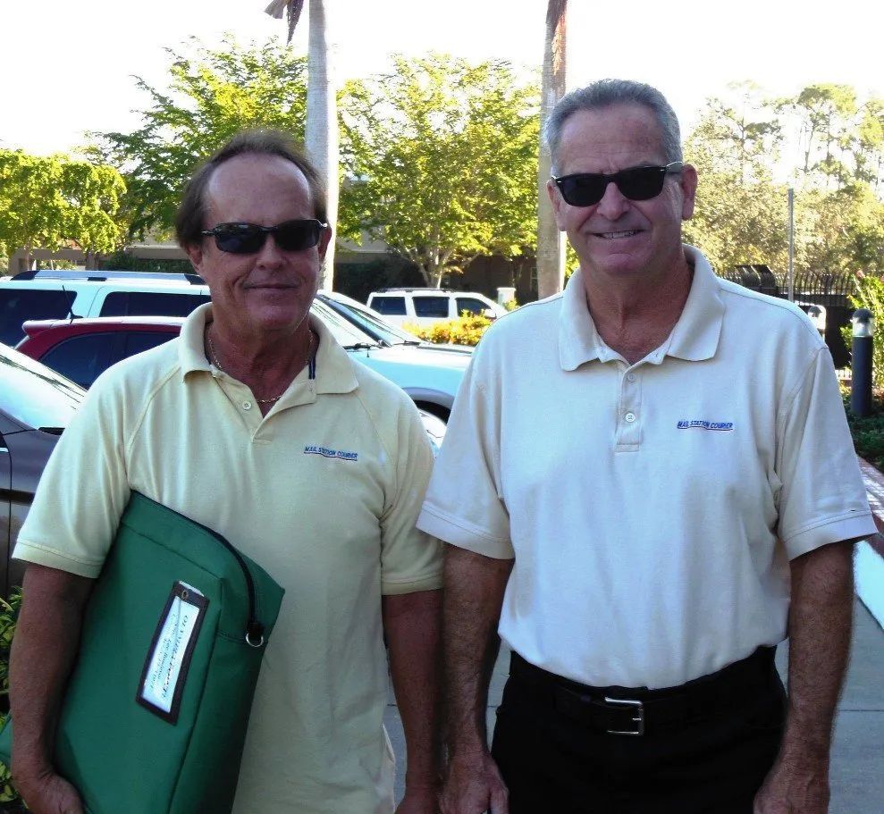 Two men in matching light-colored shirts and sunglasses stand side-by-side outdoors near parked cars and trees.