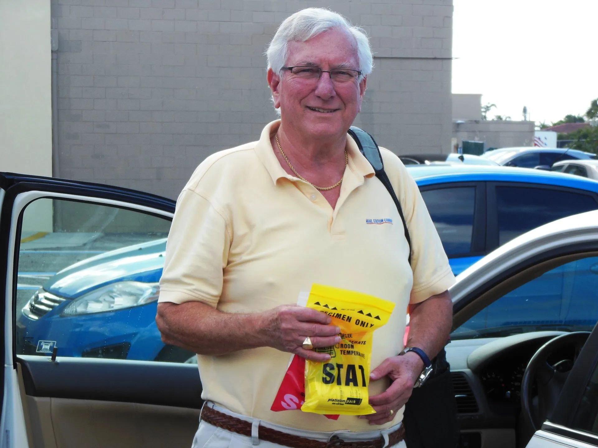Man standing near car, holding a yellow bag labeled "STATE". He wears glasses, a yellow shirt, and smiles.
