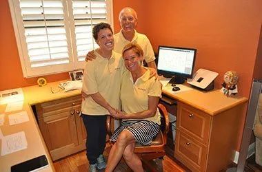 Three people in matching yellow shirts, pose in a small office with a computer and desk.