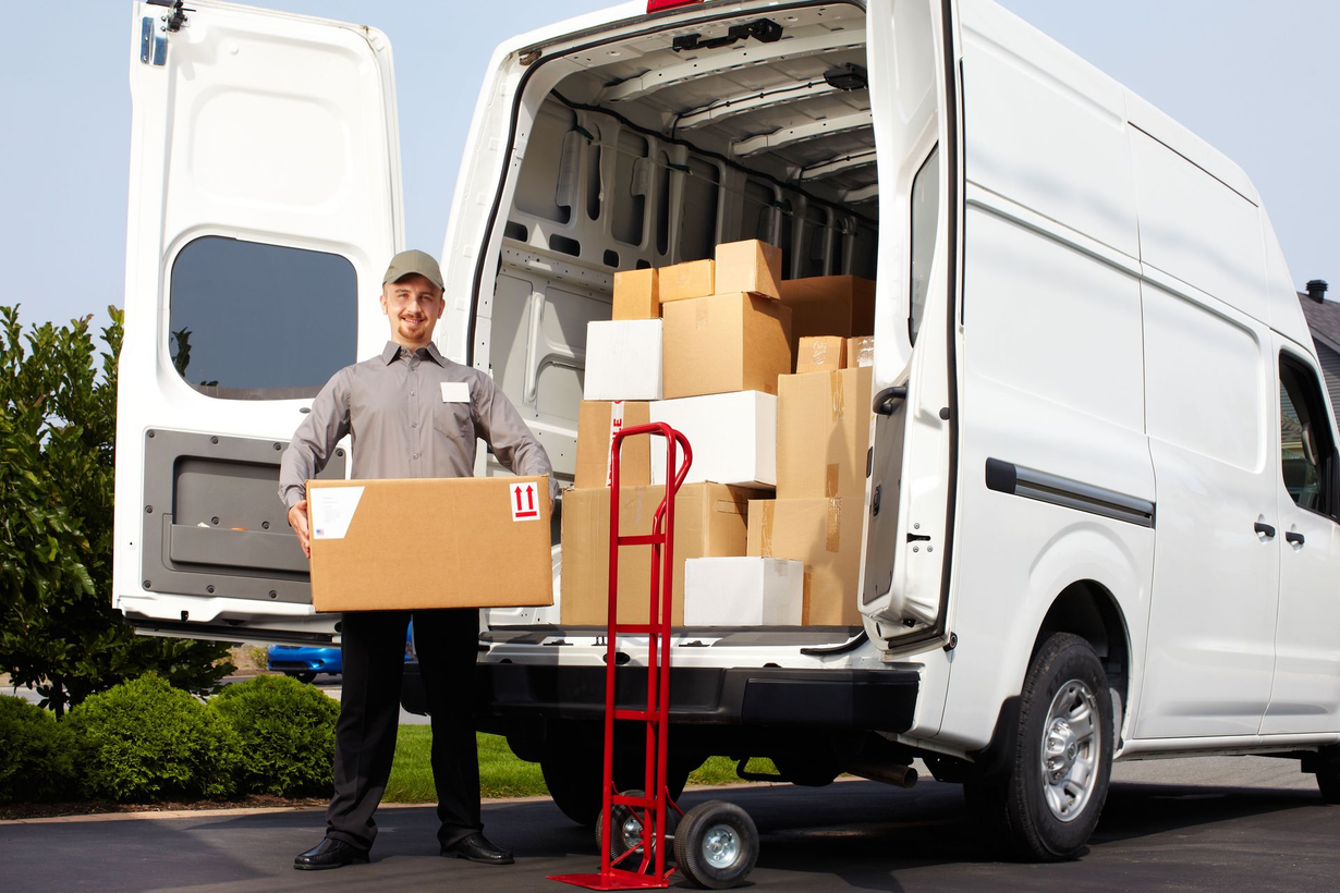 Delivery person with a cardboard box standing in front of a van filled with packages.