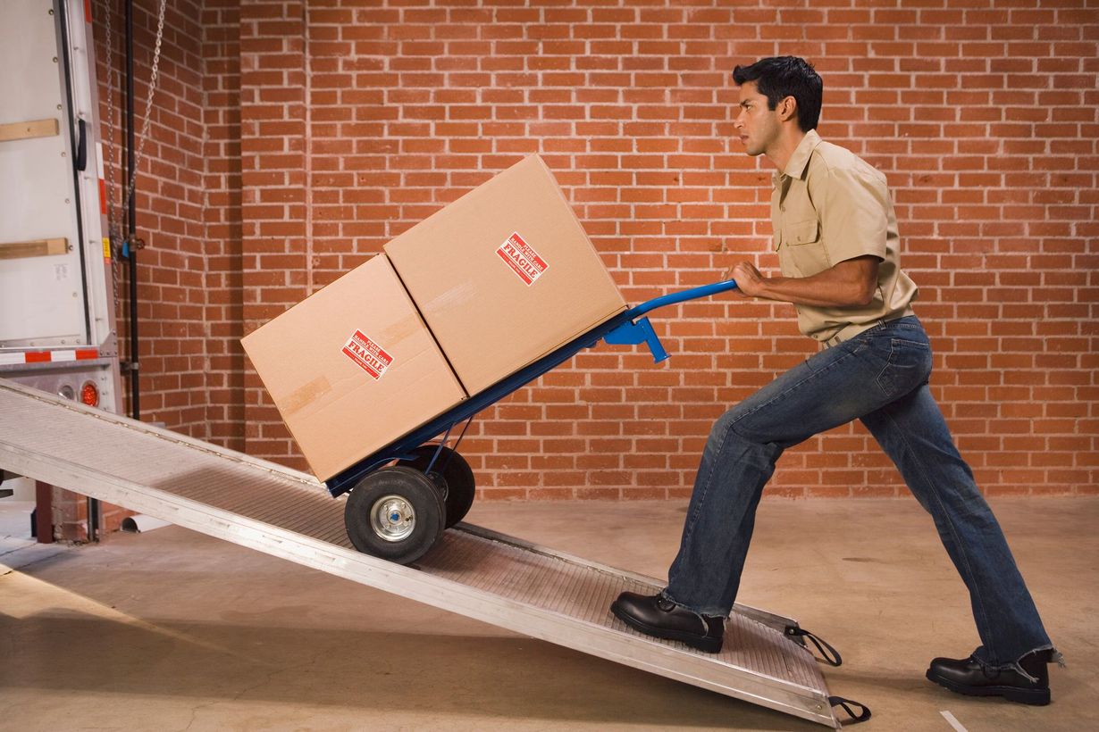 Man using a hand truck to move boxes up a ramp into a truck. Brick wall background.