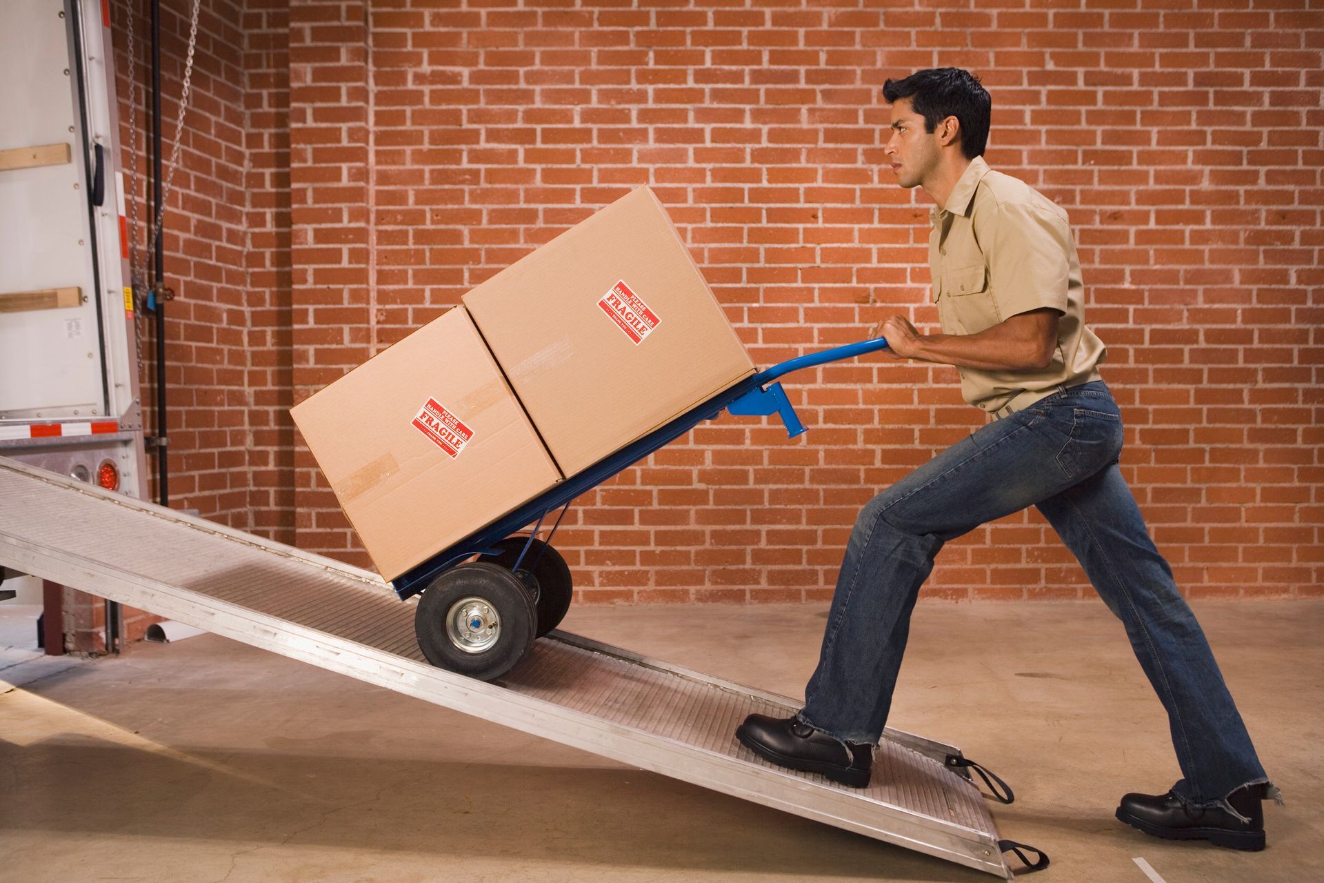 Man using a hand truck to move boxes up a ramp into a truck. Brick wall background.