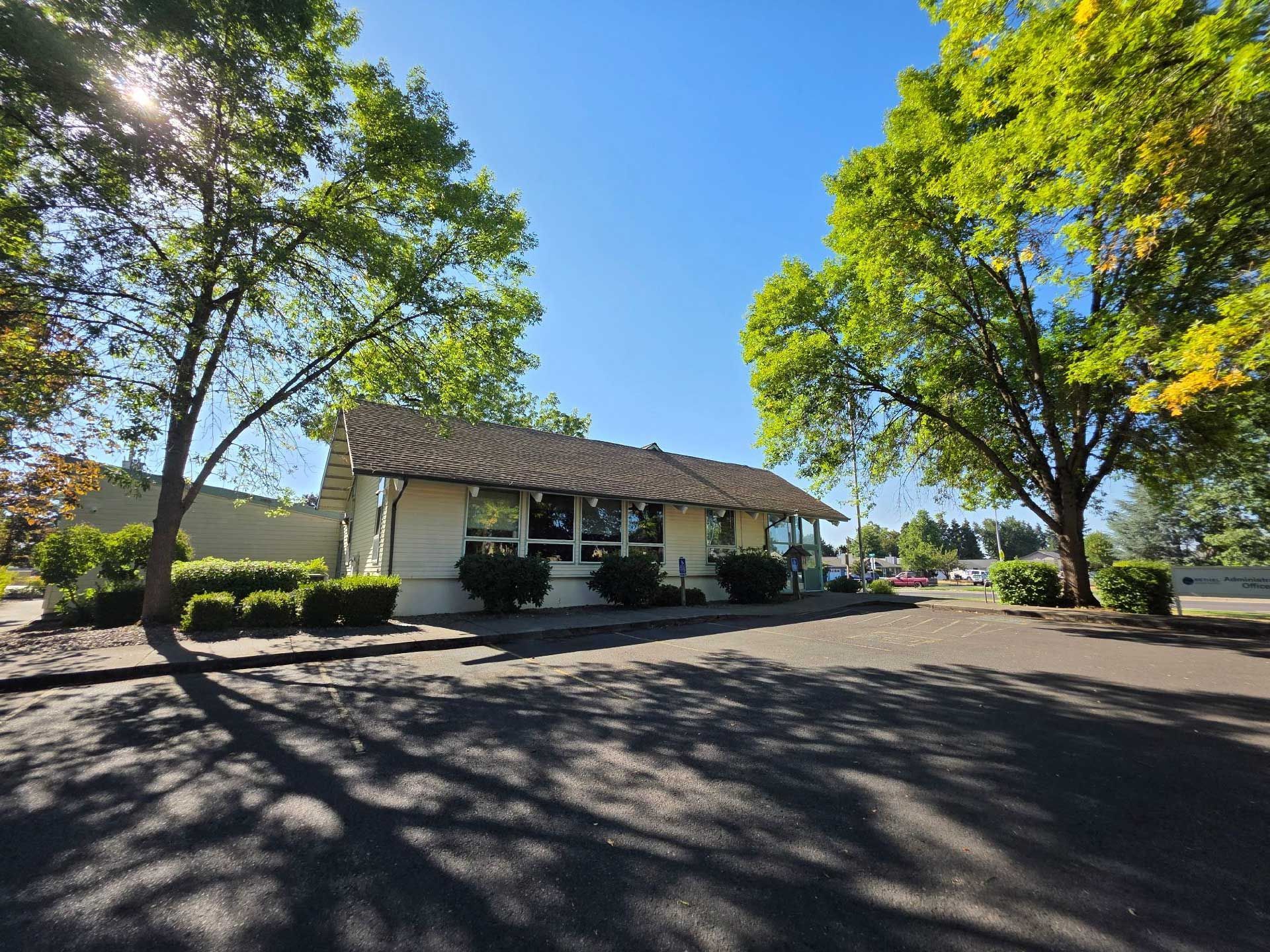 a one-story building with a tiled roof, surrounded by trees and a paved area under a bright blue sky