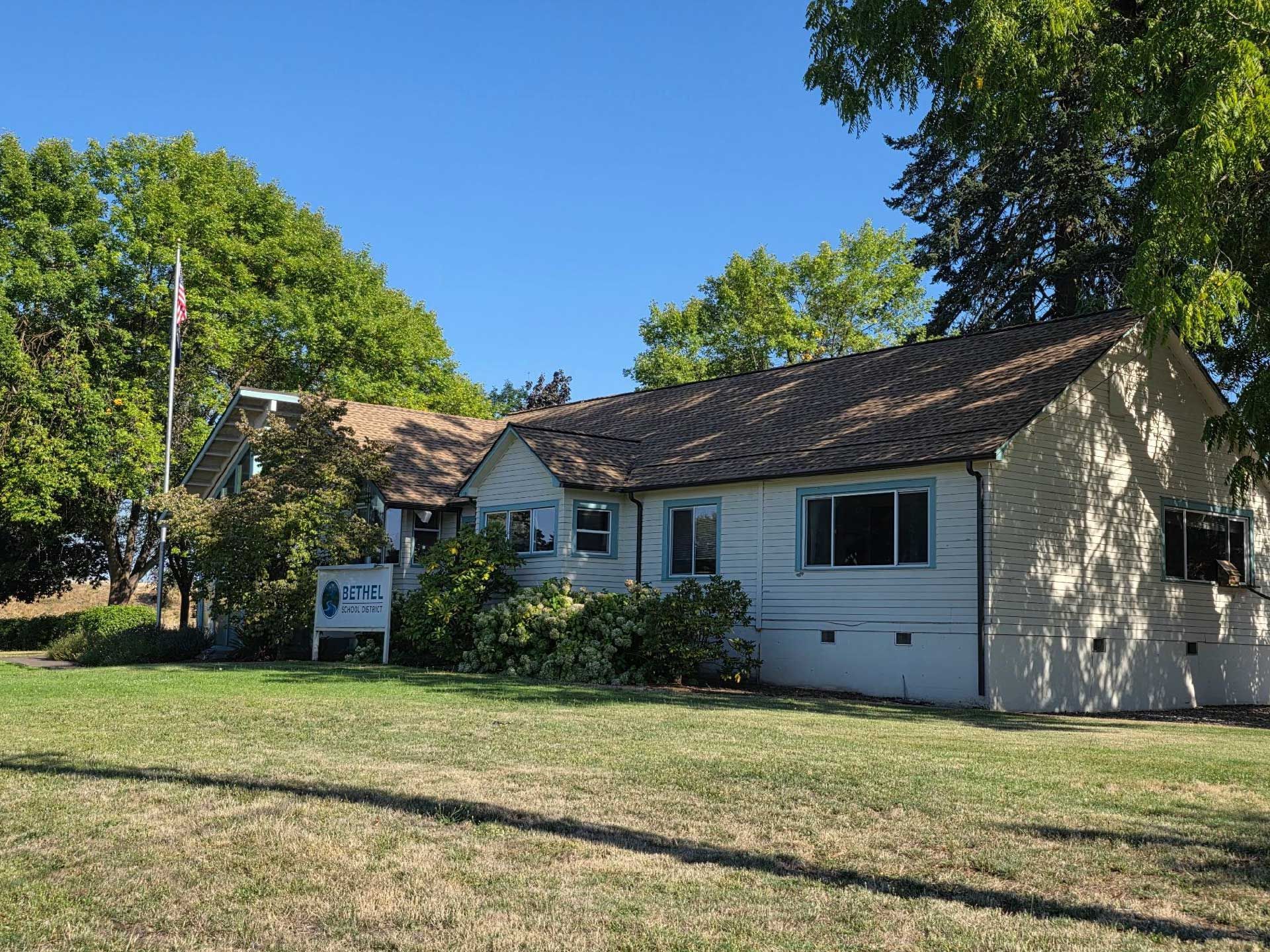 a white building with brown roof, green lawn, trees, and a sign