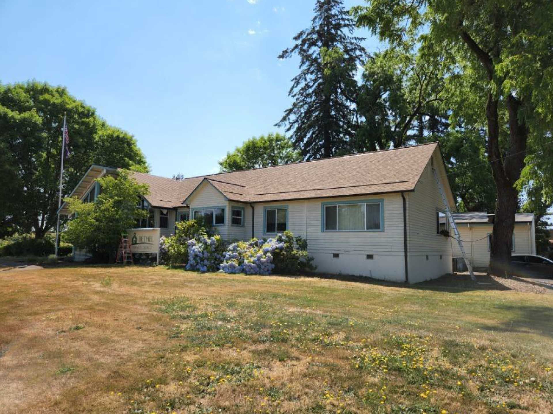 cream-colored building with brown roof and overgrown yard under a blue sky, surrounded by trees