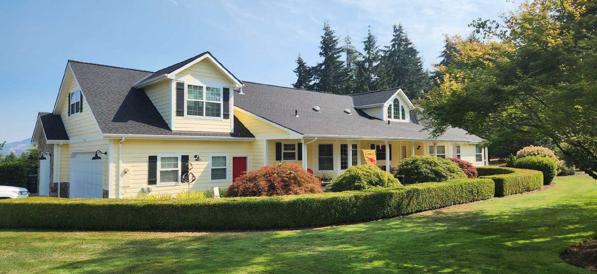 a yellow house with black shutters and red front door, surrounded by green hedges and lawn, under a blue sky