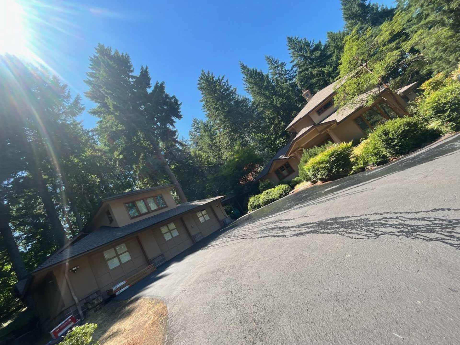 a house on a hillside surrounded by trees; sunny sky, paved driveway, brown exterior, multiple windows