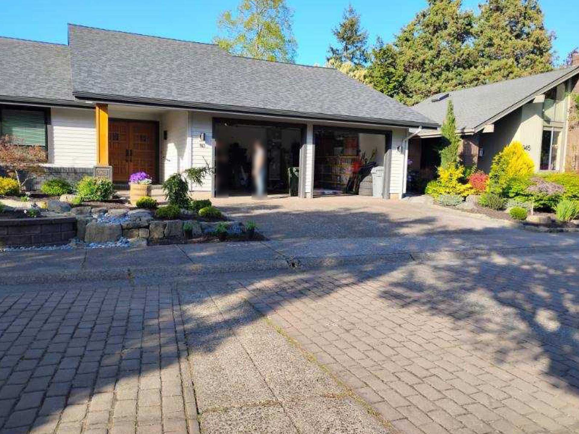 house with gray roof, brick driveway, and two-car garage, surrounded by landscaping on a sunny day