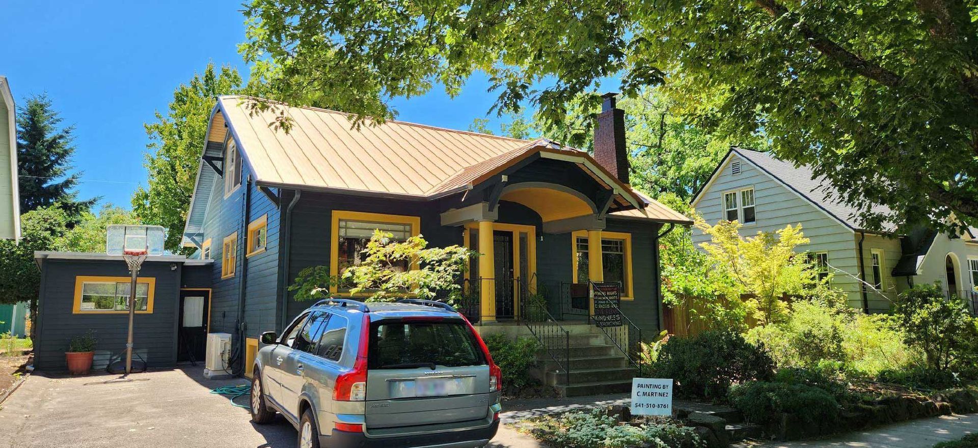 dark house with yellow trim, parked SUV, lush greenery, and clear blue sky