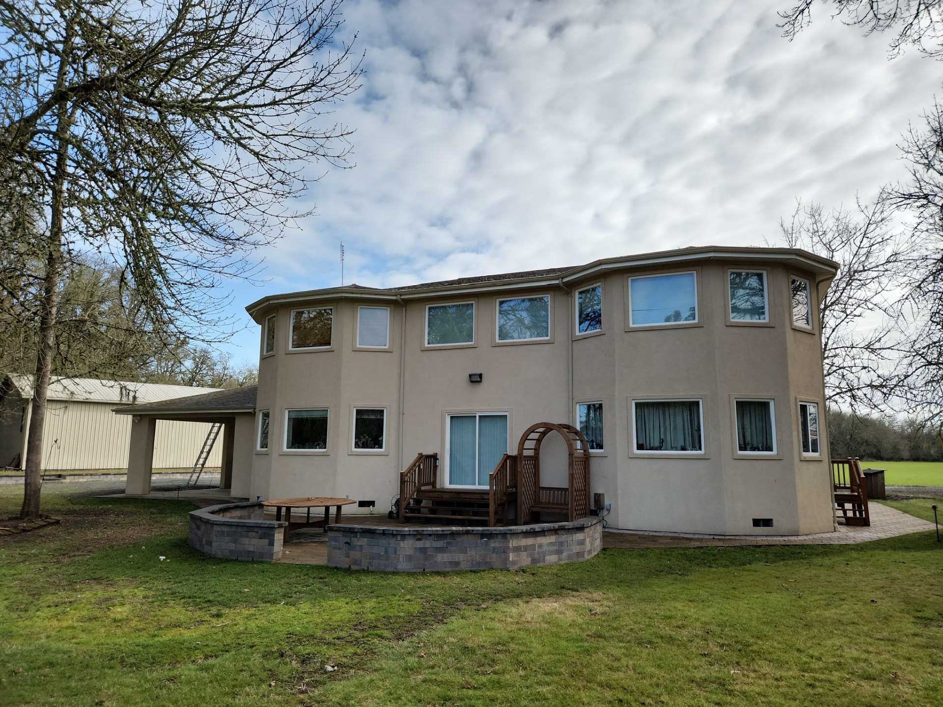 beige, curved-shape house with multiple windows, a patio, and a covered carport, set in a grassy yard under a cloudy sky
