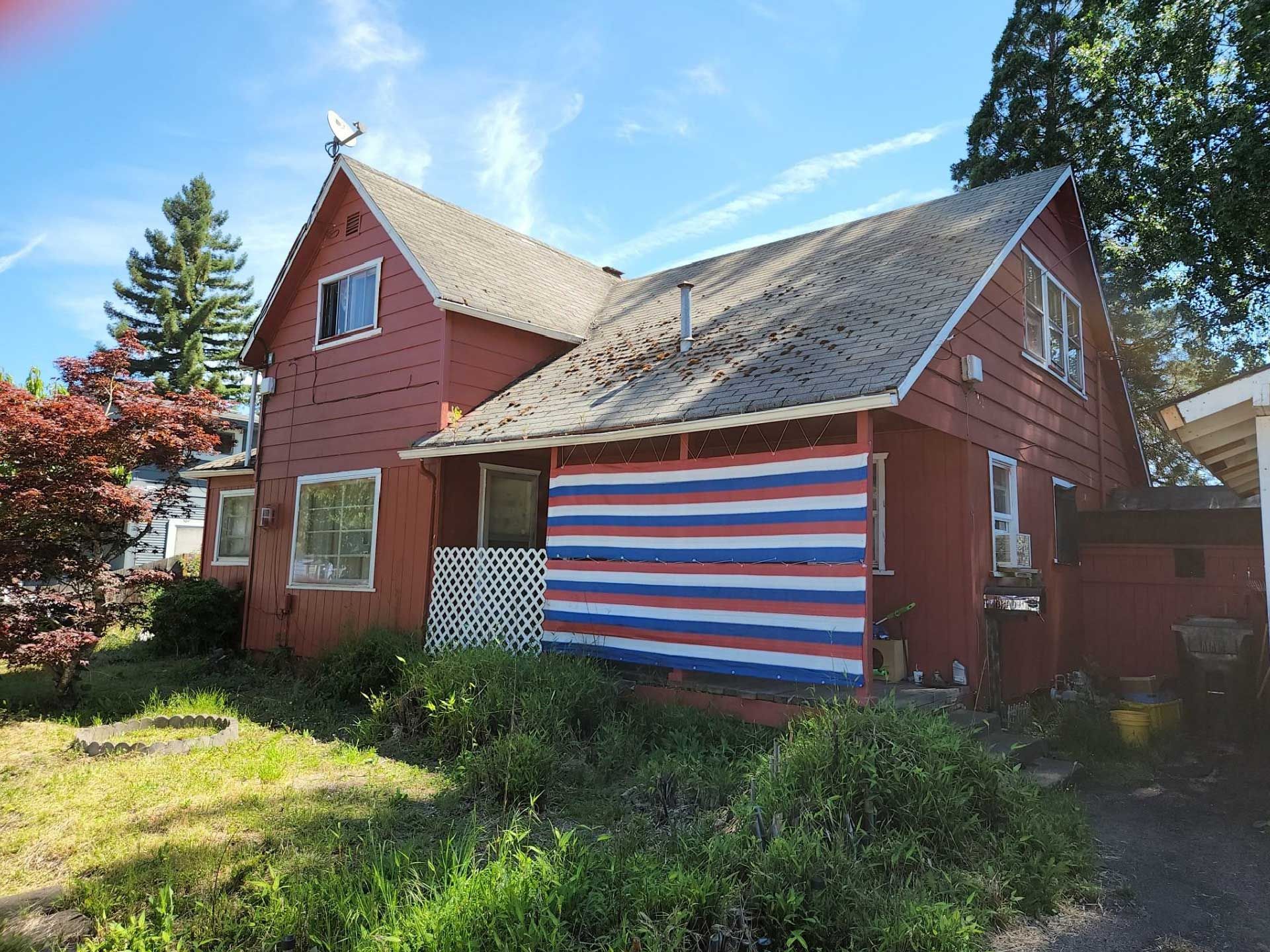 red two-story house with white, blue, and red painted facade, and overgrown yard
