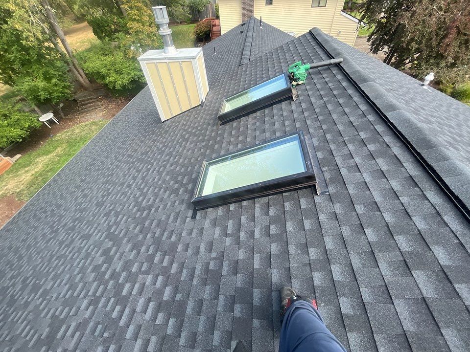 A person standing on a newly shingled roof with two skylights and a chimney. The roof is black and gray.