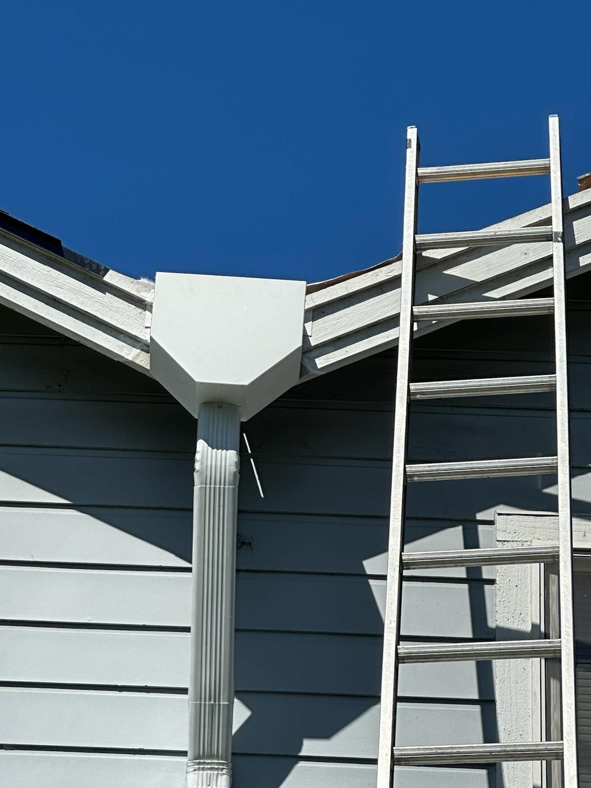 A white ladder leans against a light blue house next to a gutter and downspout under a bright blue sky.