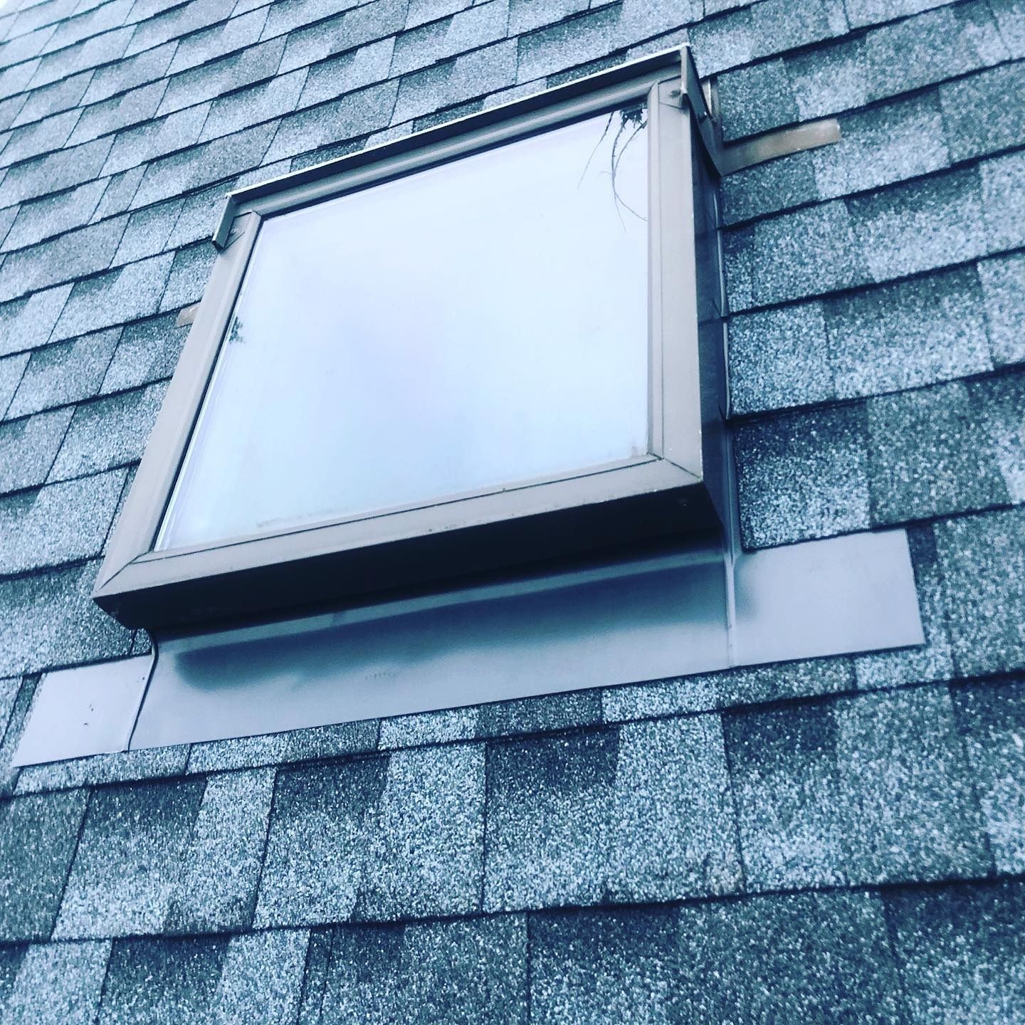 A square skylight set into a gray shingled roof, with a dark metal flashing base.