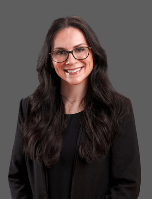 Woman with glasses, dark hair, and a smiling expression wearing a black jacket against a grey background.