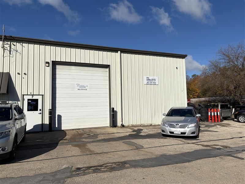 Exterior view of a white industrial building with a closed garage door. A car sits parked in front. Blue sky.