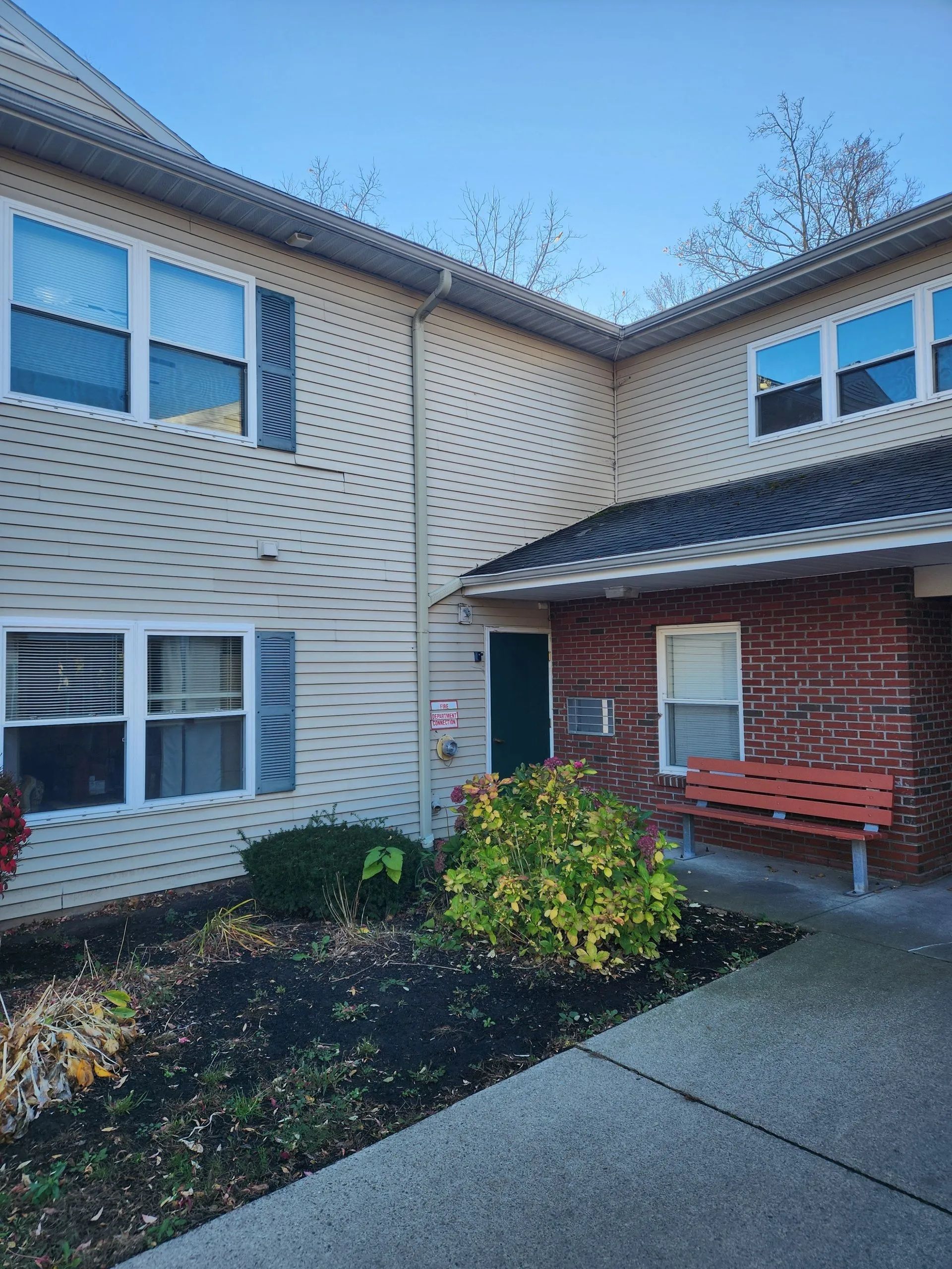Exterior of a two-story apartment building with tan siding, brick entryway, and a bench.
