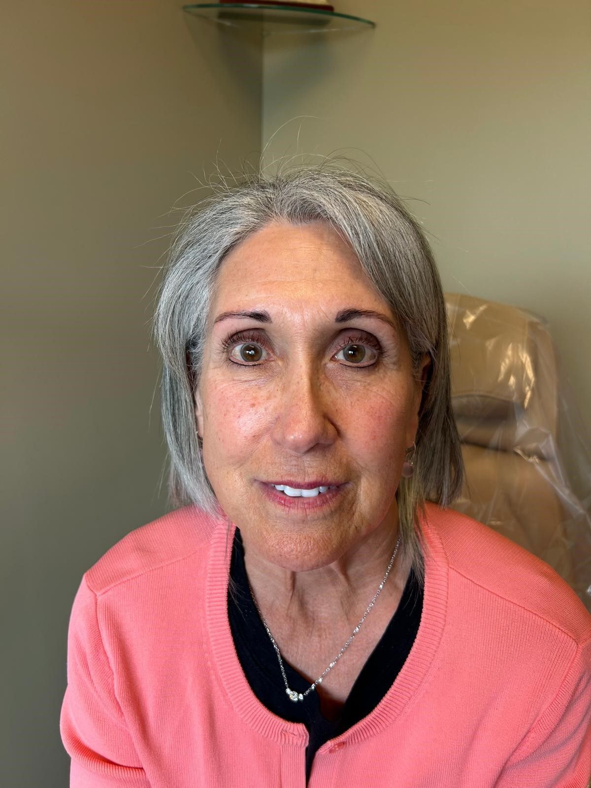 A woman with gray hair is wearing a pink sweater and sitting in a chair.