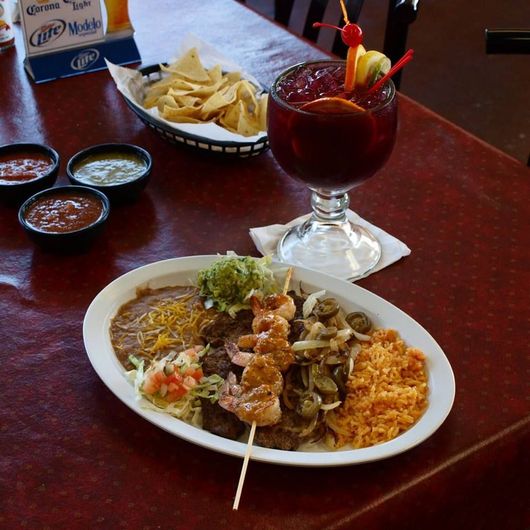 Mexican food platter with shrimp skewers, beans, rice, and salsa, plus chips and a drink.