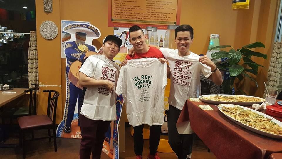 Three people holding up a t-shirt at a restaurant; one is Asian, and two are men.