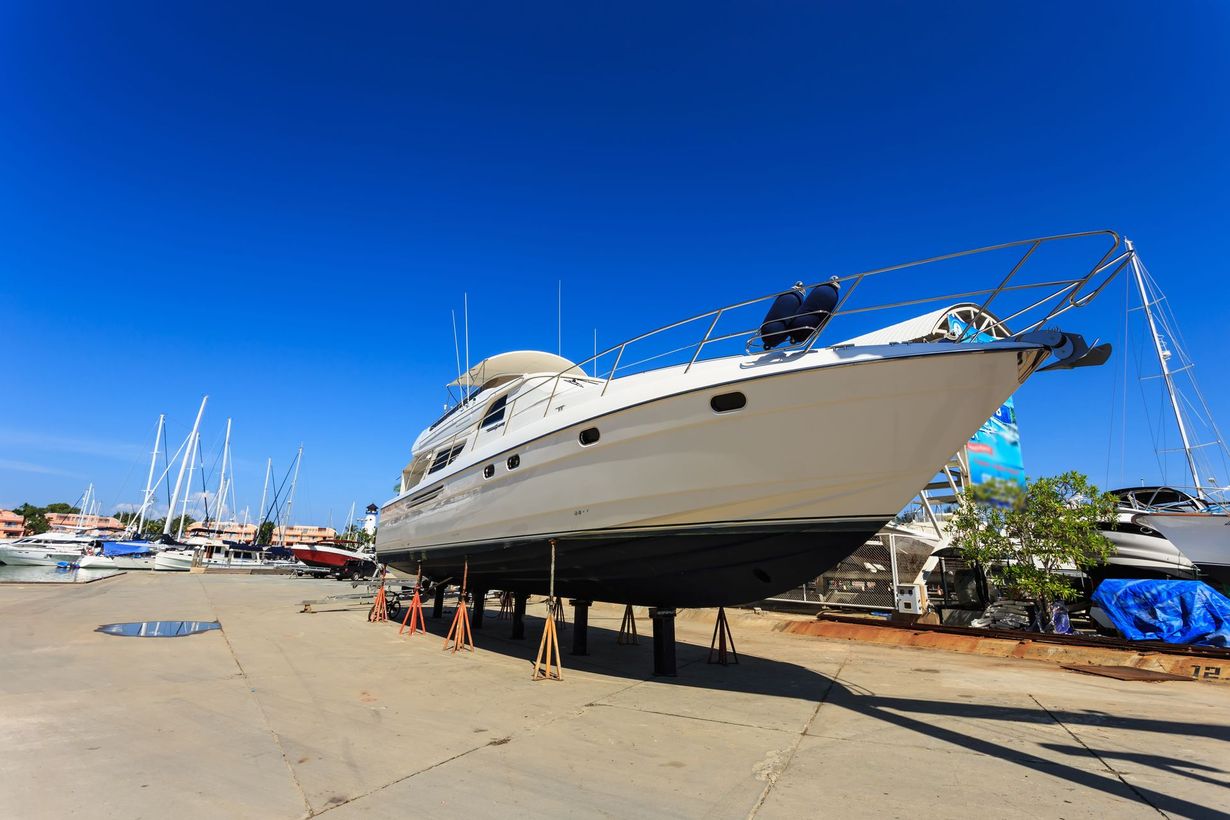 A large, white yacht propped up on supports in a sunny boatyard against a clear blue sky.